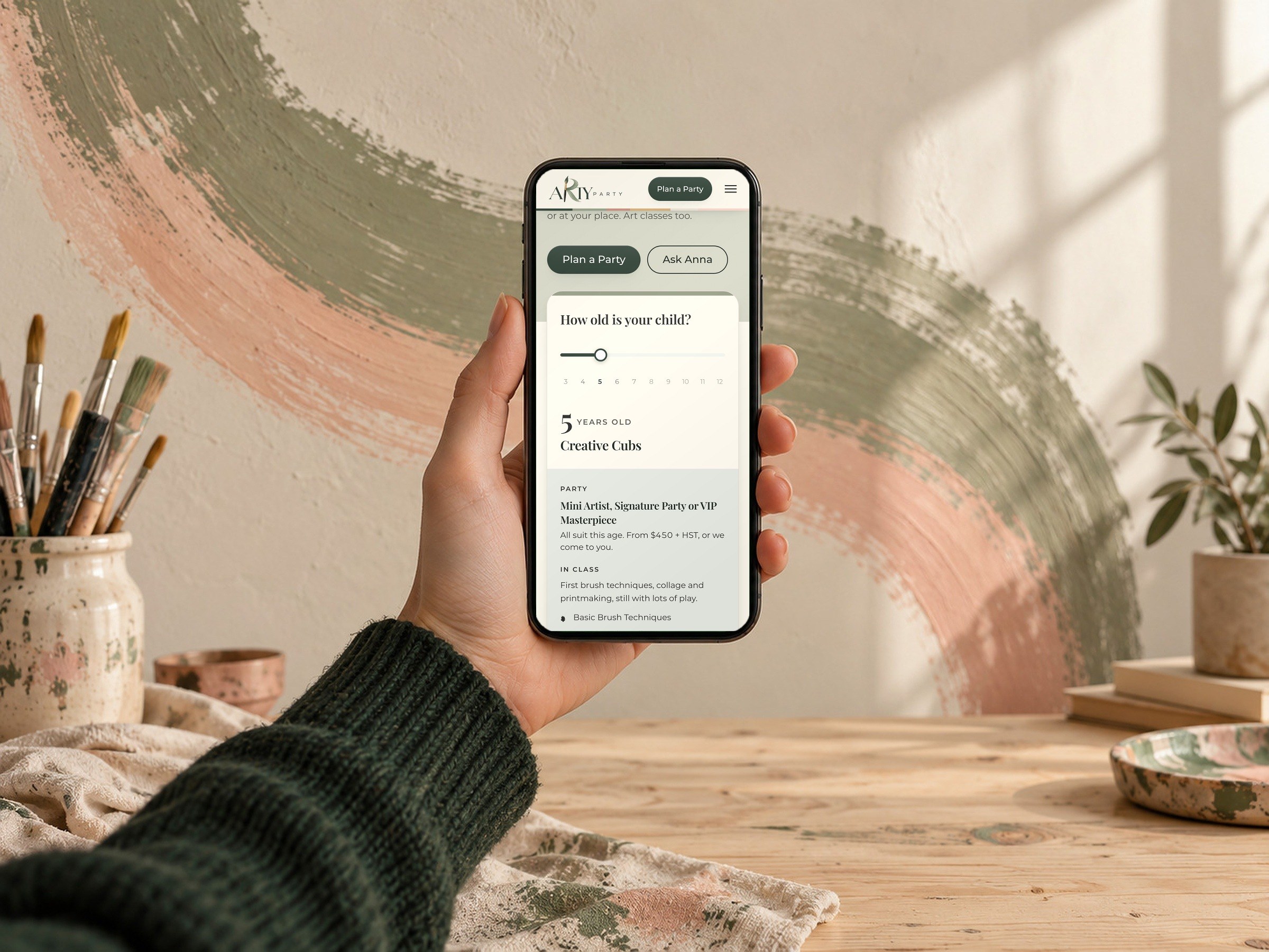 The age finder from the Arty Party home page on a phone, held in front of a painted brushstroke.